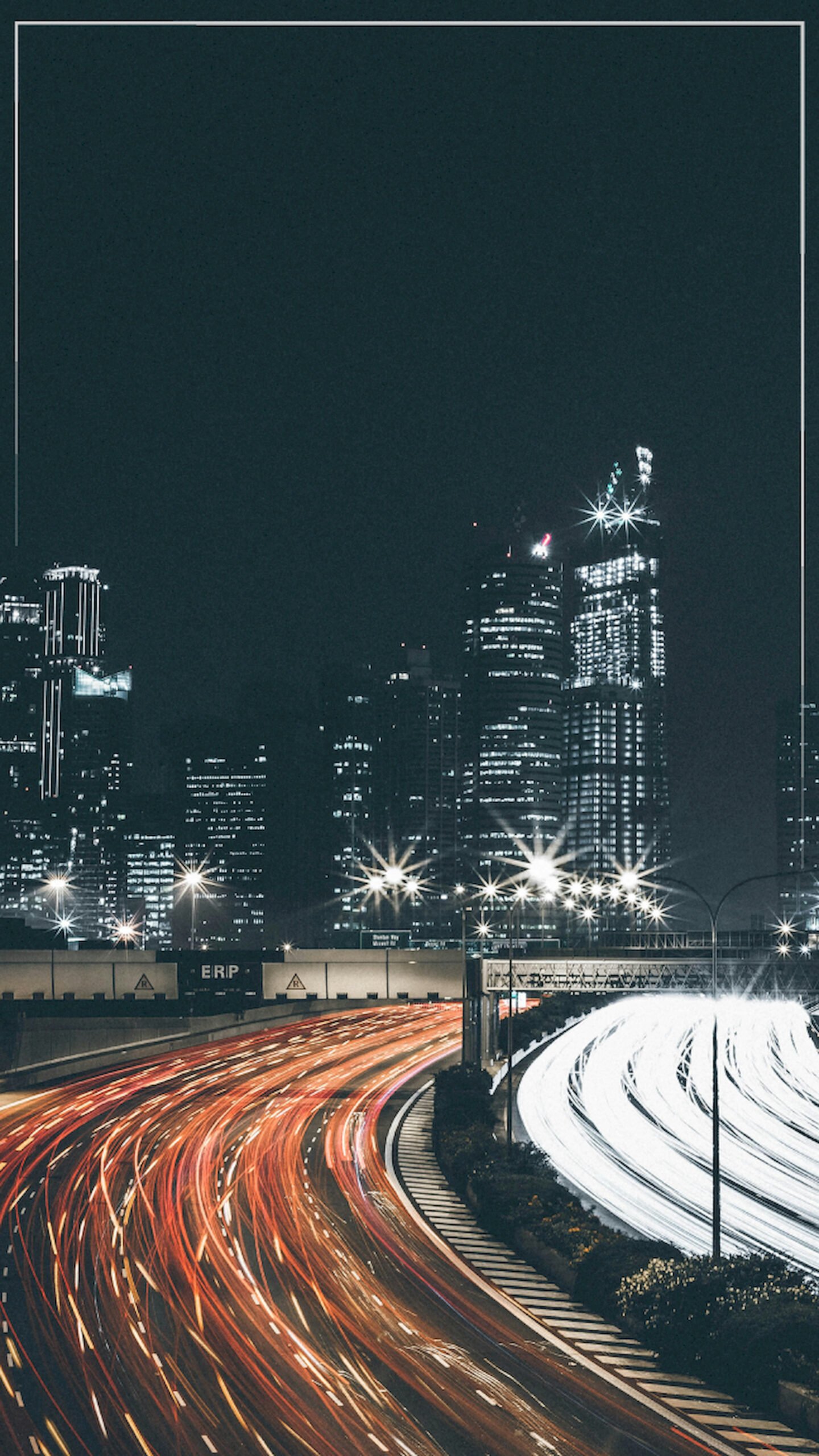 "Vertical long-exposure night photography of a city highway showing curved red and white light trails from traffic leading towards tall illuminated skyscrapers."
