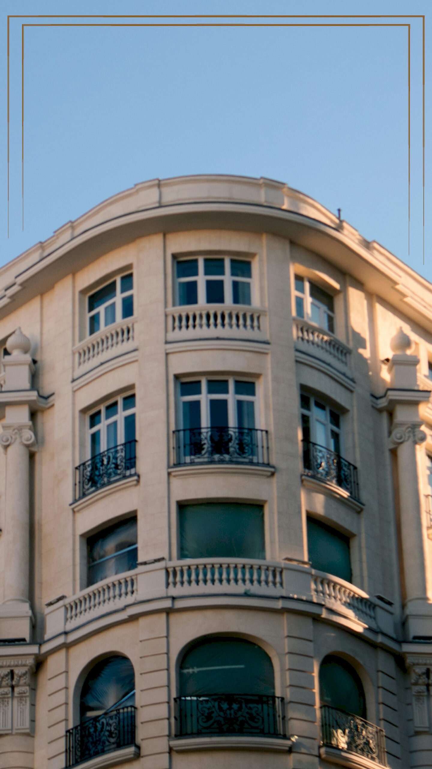"Low angle view of a classic European corner building with beige stone, balconies, and arched windows under a blue sky, vertical 4K wallpaper."