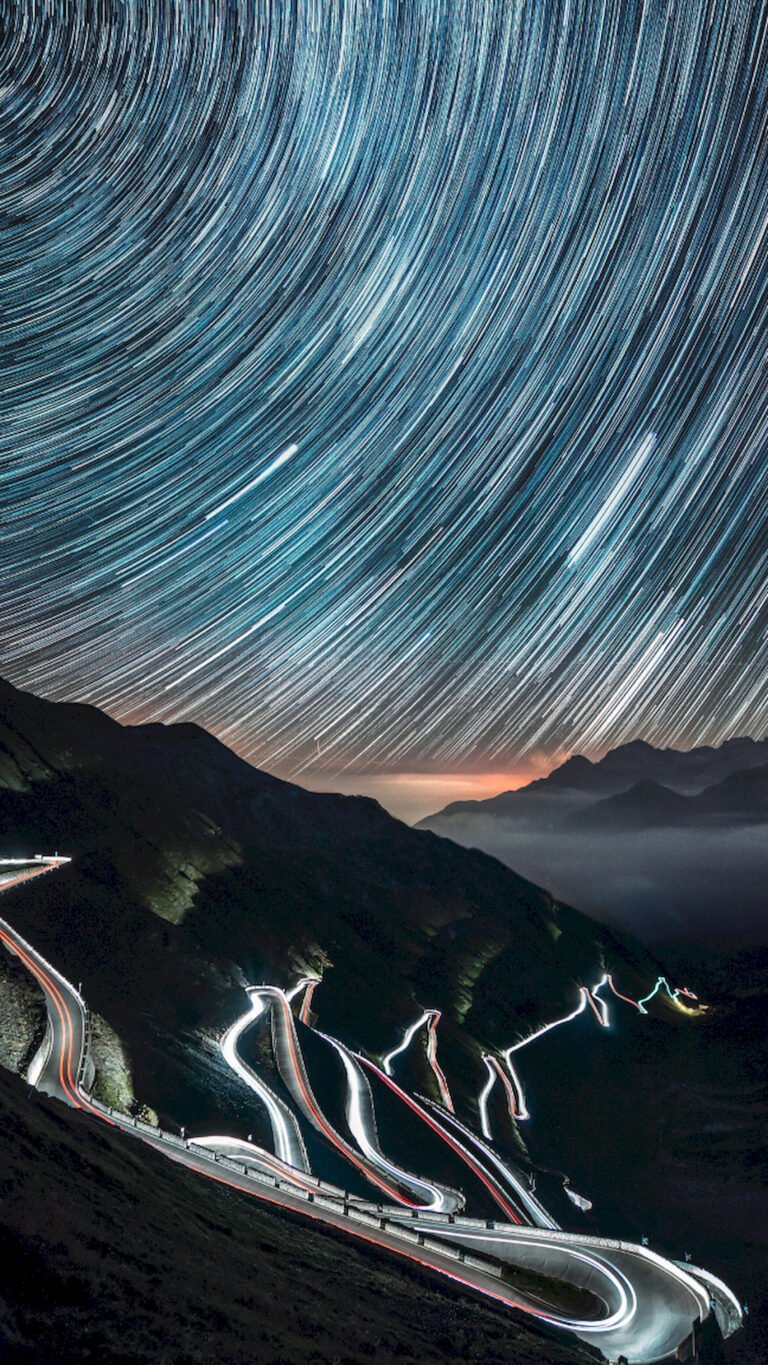 "Vertical long-exposure night photography showing circular star trails in the sky over a winding mountain road illuminated by car light trails."