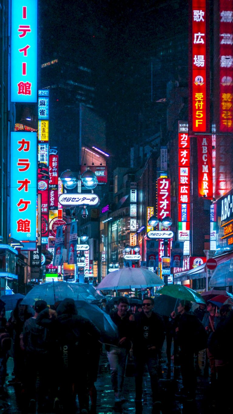 "Vertical night photo of a busy street in Tokyo, Japan, filled with bright neon signs, rain, and people walking with clear umbrellas."