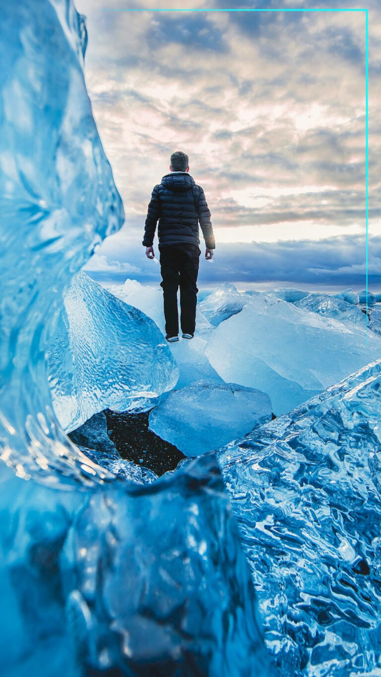 "Man standing on large blue ice blocks on a beach in Iceland, looking out at a cloudy sky during sunset, 4K phone wallpaper."