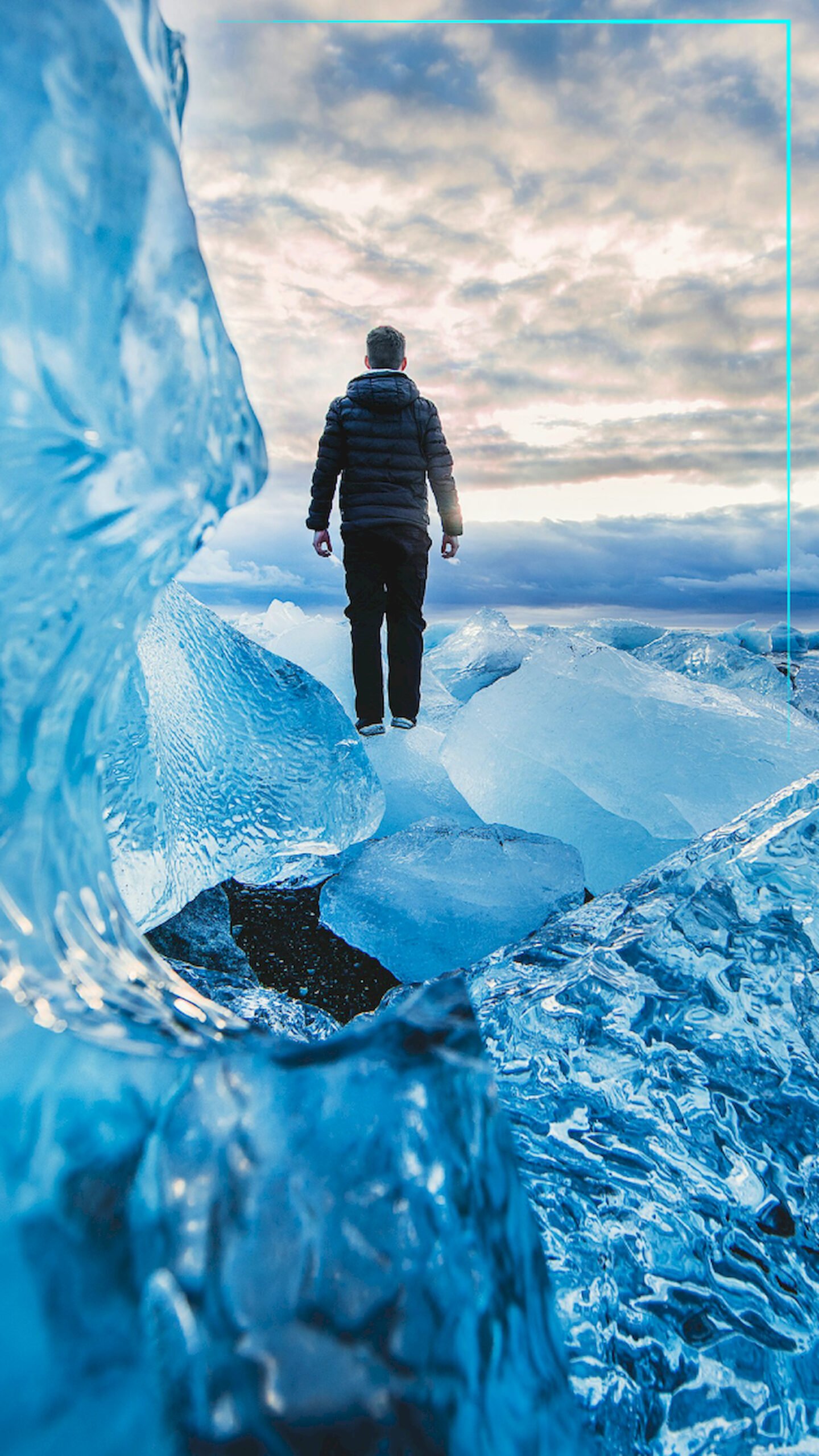 "Man standing on large blue ice blocks on a beach in Iceland, looking out at a cloudy sky during sunset, 4K phone wallpaper."