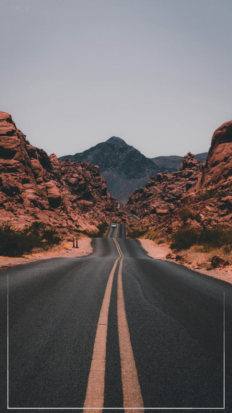 "Vertical view of a long asphalt road stretching through red desert rock formations towards a dark mountain under a clear sky."