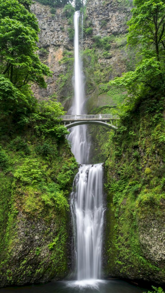 A towering waterfall flowing under an arch bridge surrounded by lush green trees and mossy rock cliffs.