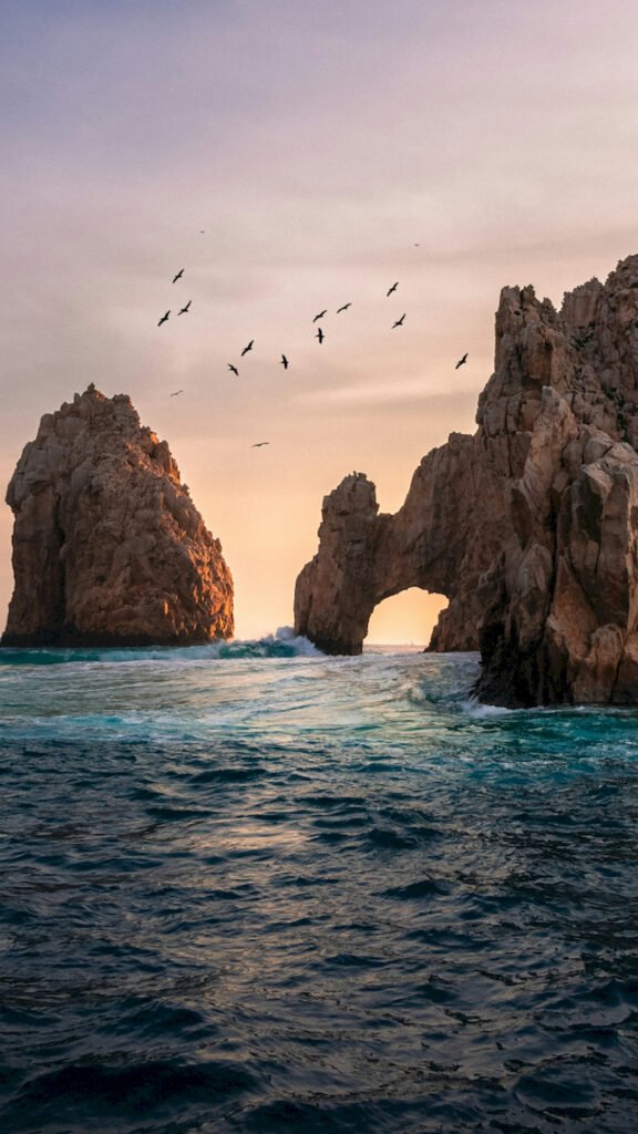 Scenic view of a natural rock arch in the ocean at sunset with a flock of birds flying over crashing waves.