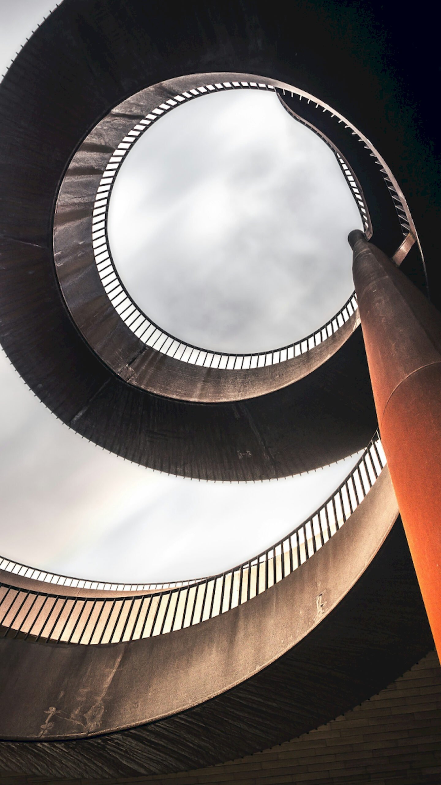 "Worm's-eye view looking up at a concrete spiral ramp with a cloudy sky in the center, vertical 4K wallpaper."