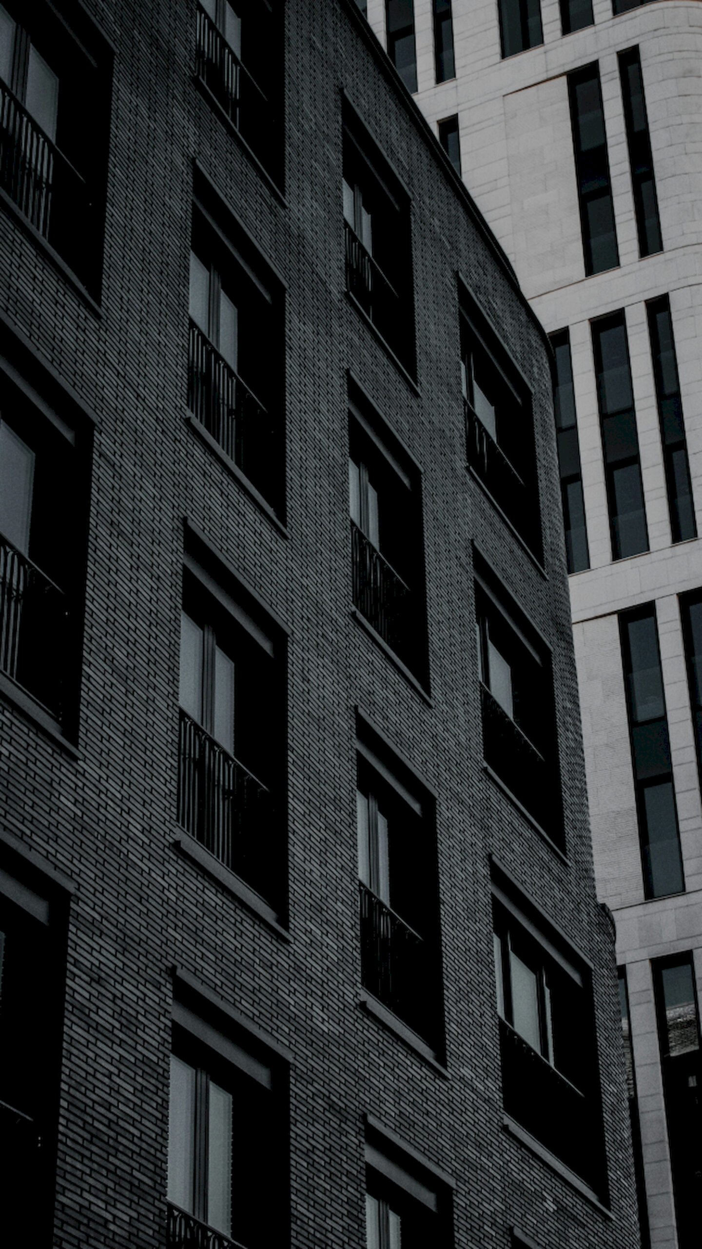 "Vertical low-angle view of a dark grey brick apartment building with balconies standing next to a modern white stone high-rise building."