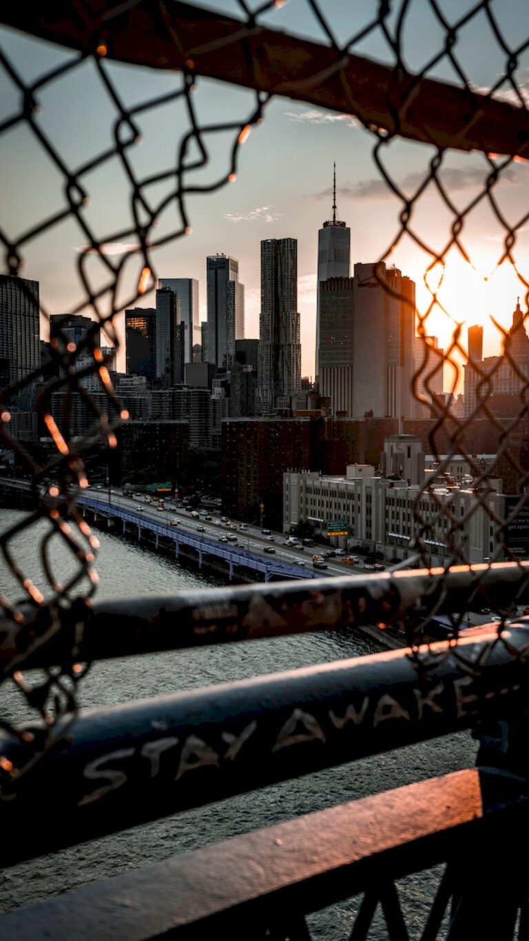 "Vertical urban photography of New York City skyline and One World Trade Center at sunset seen through a chain-link fence with 'STAY AWAKE' written on the railing."