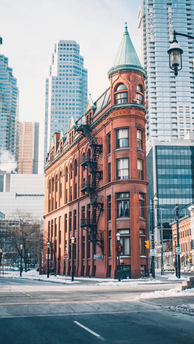 "Vertical winter view of the historic red brick Gooderham Flatiron building in Toronto with a green roof, standing in contrast to modern glass skyscrapers on a snowy street."