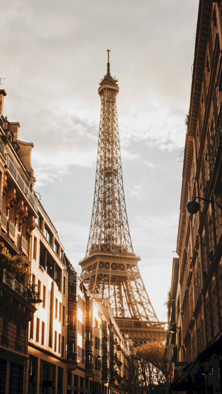 "Vertical street view of the Eiffel Tower in Paris, framed by residential buildings with balconies under a warm cloudy sky."