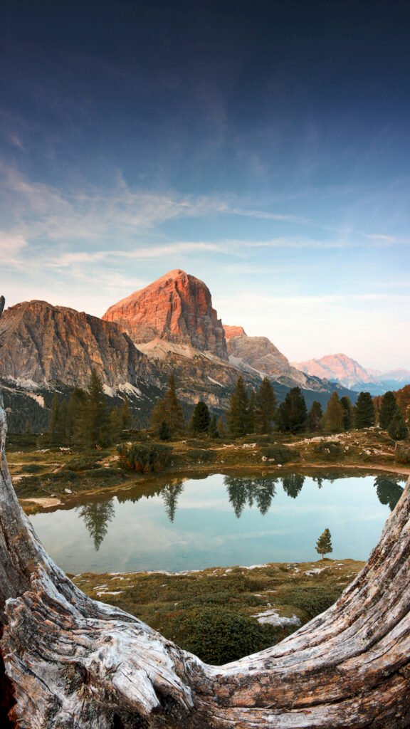 A glowing mountain peak at sunset reflected in a calm lake, framed by a textured weathered tree log in the foreground.
