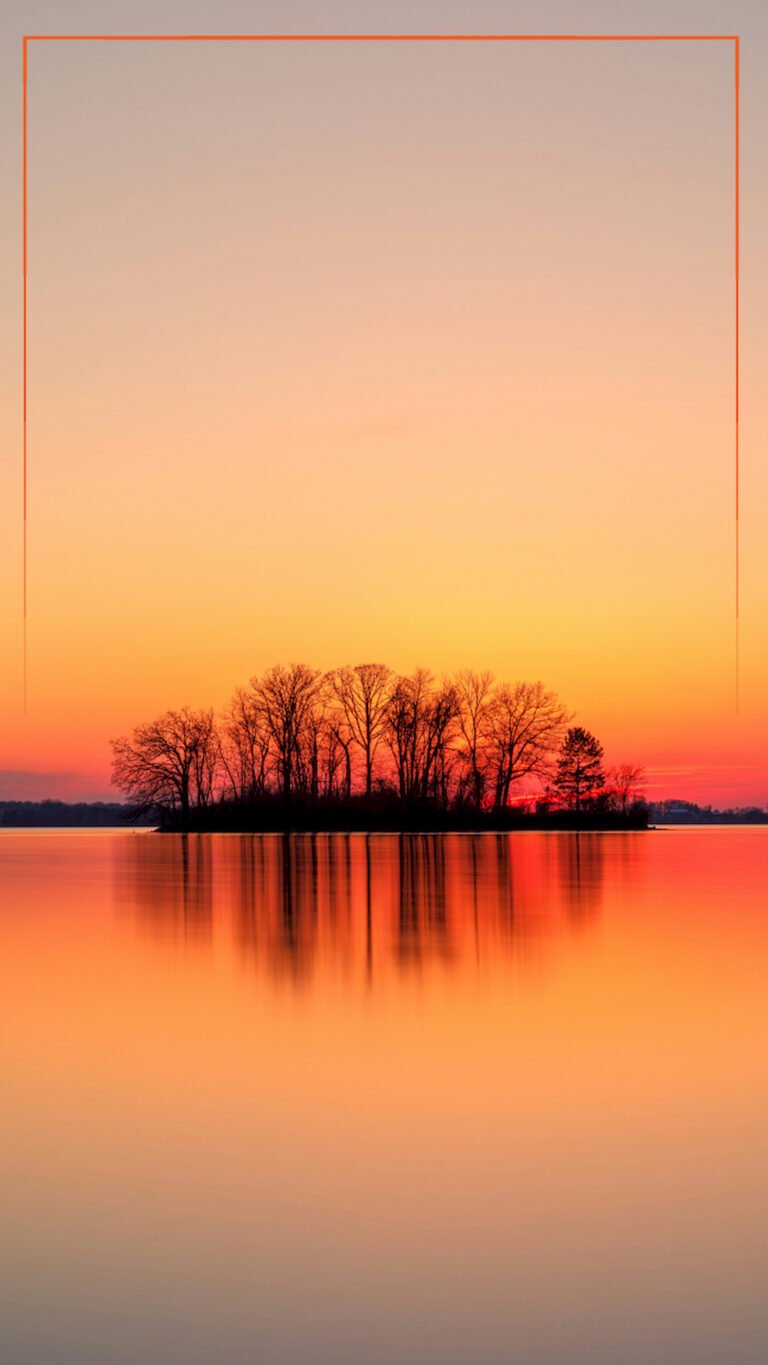 Silhouette of a small island with trees centered on a calm lake against a vibrant orange and yellow sunset sky with a perfect water reflection.