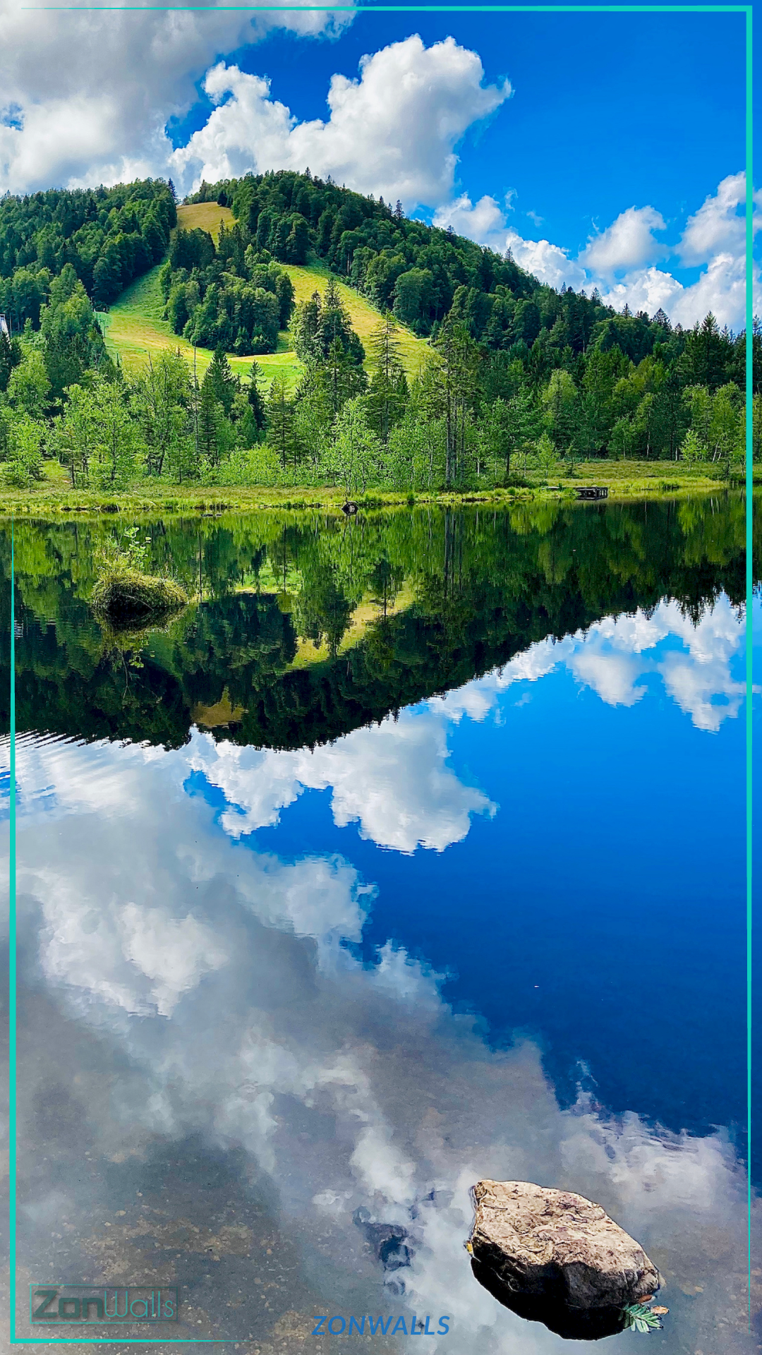Vertical nature wallpaper featuring a crystal-clear blue lake mirroring a lush green forest hill and fluffy white clouds, with a single rock submerged in the foreground.