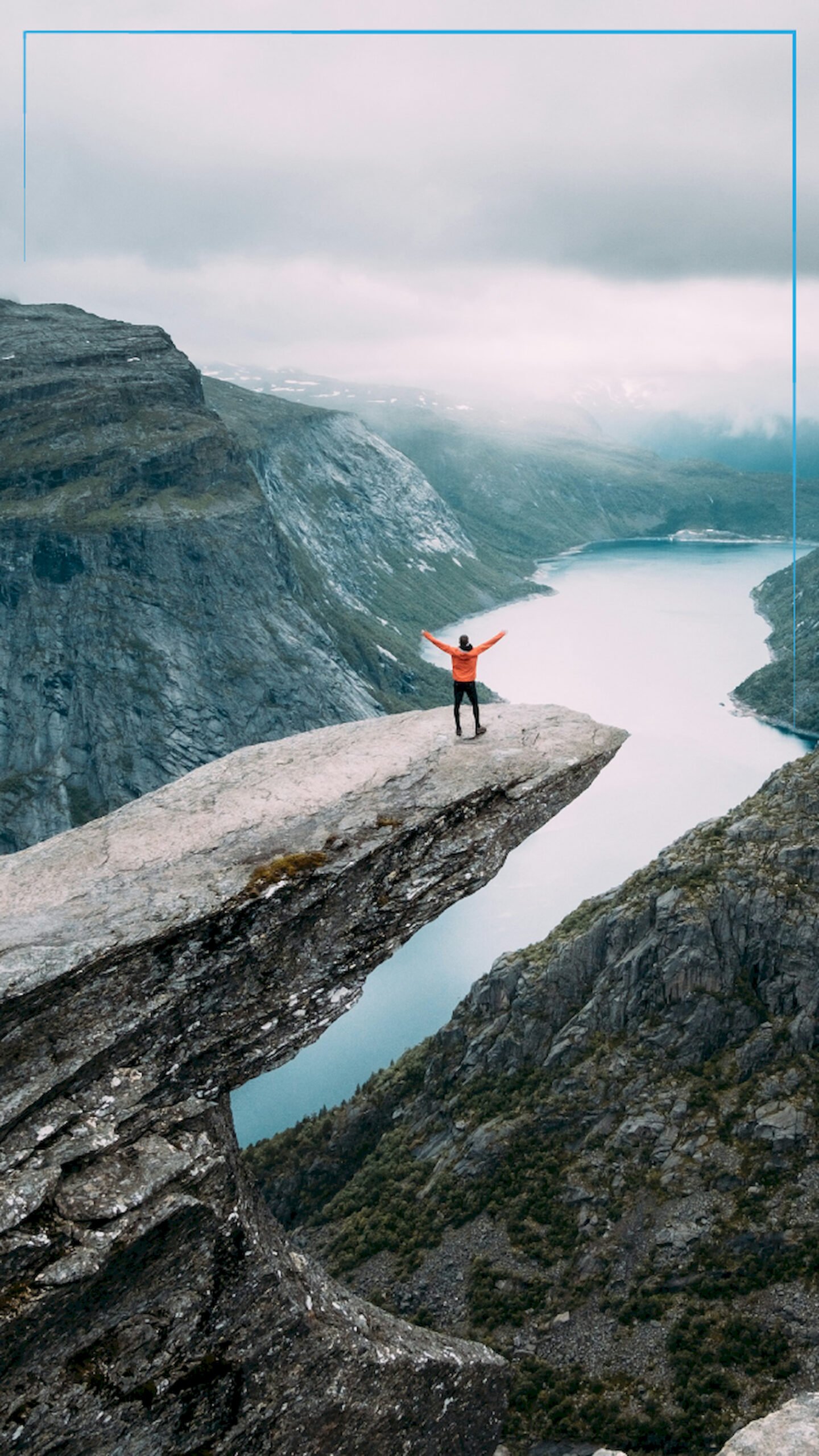 "Man standing with arms raised on the edge of Trolltunga cliff in Norway, overlooking a lake and mountains on a cloudy day."